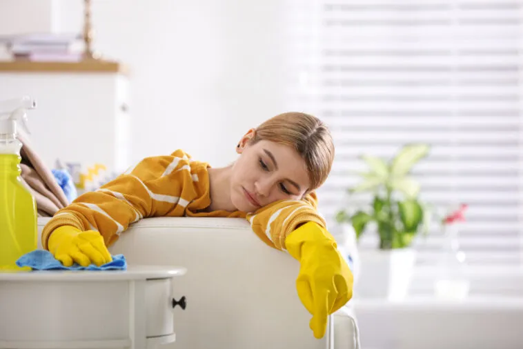 woman in yellow cleaning gloves looks exhausted while cleaning her home. She is resting her head on the back of the couch, surrounded by cleaning supplies.
