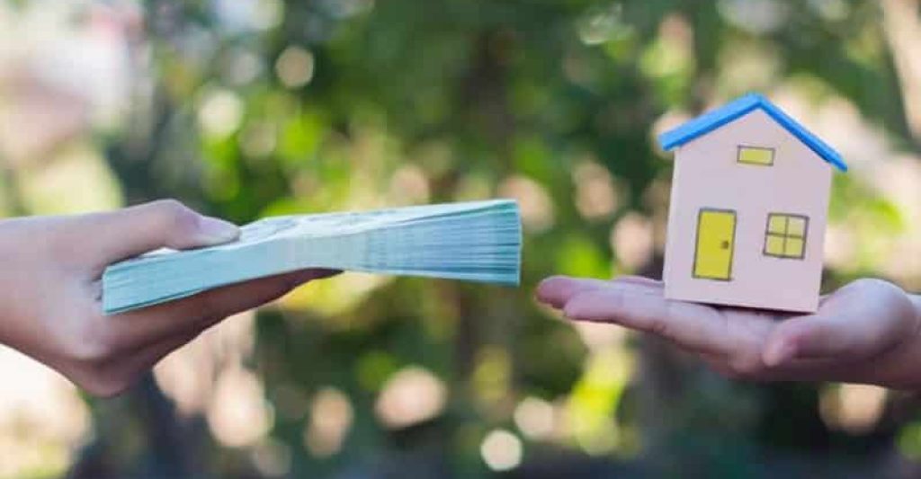  A close-up image of hands exchanging a bundle of cash for a small house model, symbolizing property purchase or investment.