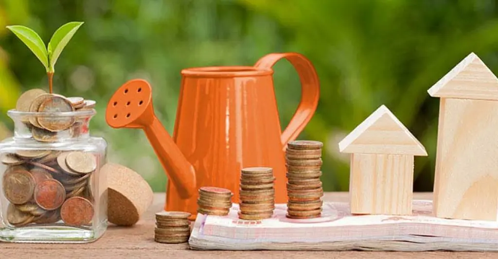 A conceptual image showing home investment growth with coins stacked, a watering can, and wooden house models on a wooden surface.