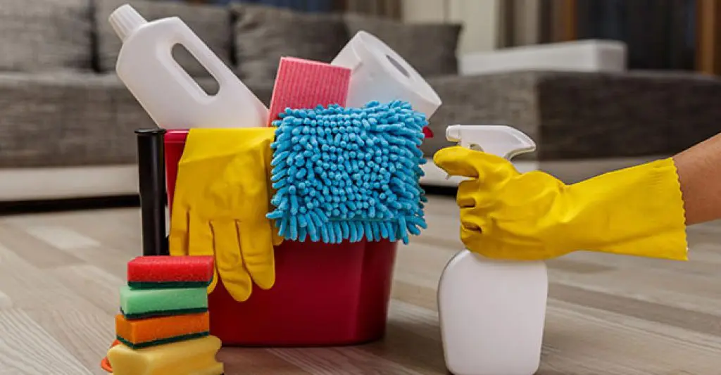 A red bucket with cleaning supplies, gloves, and sponges placed on a wooden floor, ready for household cleaning.