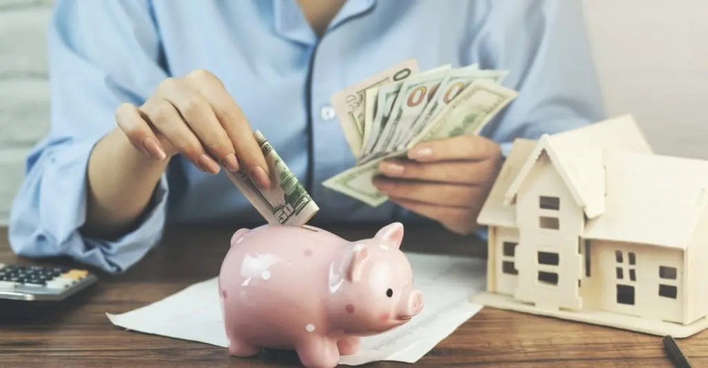 A person inserting cash into a pink piggy bank beside a wooden house model, representing home savings or budgeting.