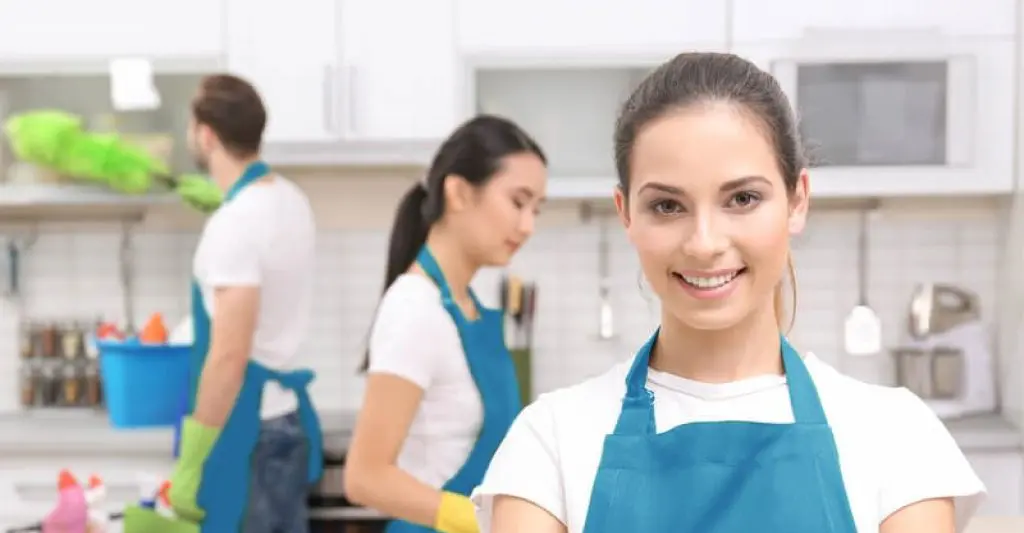 Smiling female cleaner in focus with a team working in the background inside a modern kitchen in Sydney, wearing blue aprons and gloves.
