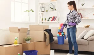 A woman stands with cleaning tools in a living room filled with moving boxes, preparing for a move-in clean.