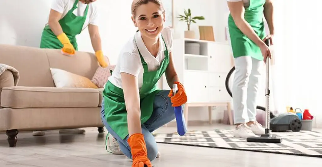 A cheerful cleaning team in green aprons scrubbing the floor and vacuuming an apartment interior.