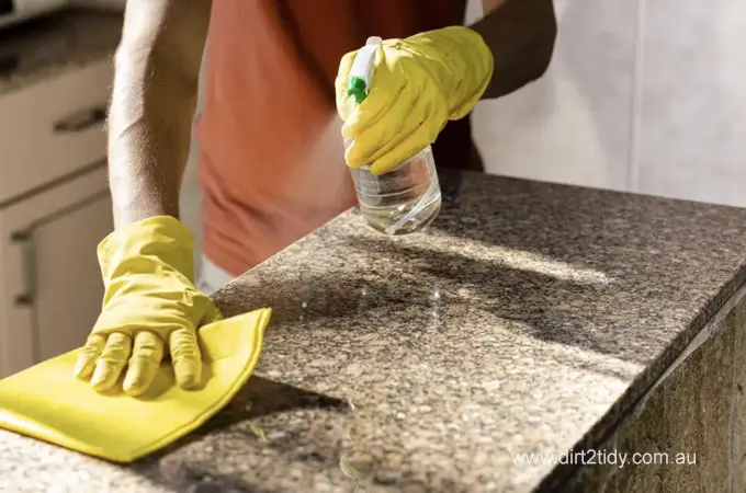 Need to do a Deep Clean for End of Lease Cleaning 3 Cleaner spraying disinfectant on a granite kitchen countertop while wiping with a yellow cloth.
