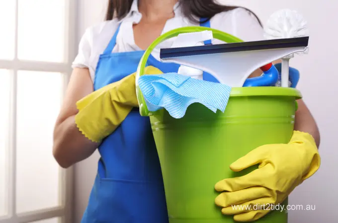 Smiling cleaning lady holding a bucket of supplies with a team of professional cleaners in the background – representing Dirt2Tidy’s expert cleaning crew.