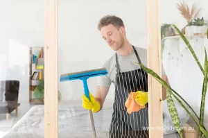 Male cleaner wearing gloves and apron using a squeegee to clean glass – representing professional window cleaning services by Dirt2Tidy.