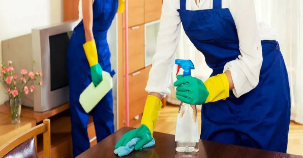 Two professional cleaners in uniforms disinfecting and wiping down furniture surfaces.