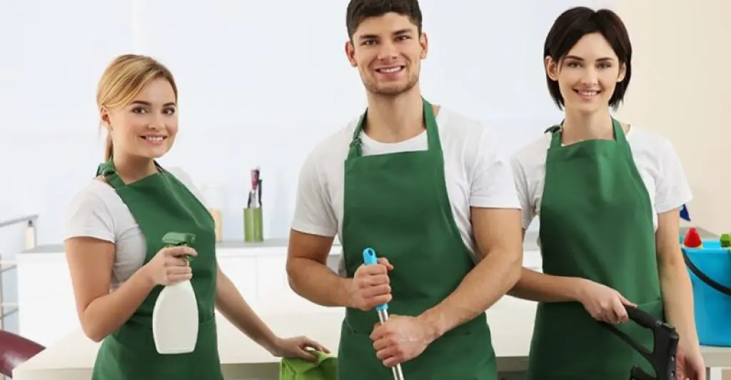 Three smiling professional cleaners wearing green aprons holding cleaning tools – ready to provide expert cleaning services with a friendly attitude.

