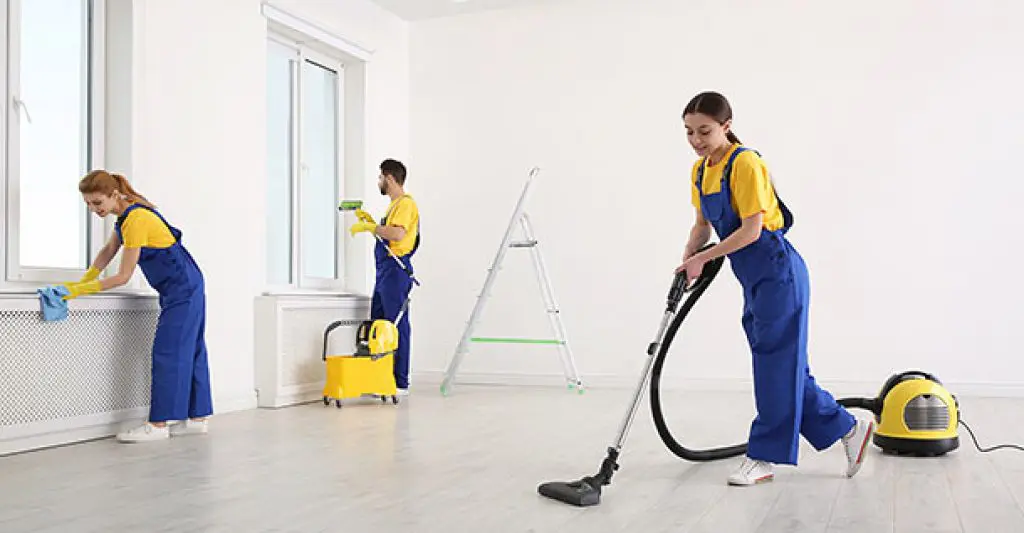 A group of professional cleaners in blue and yellow uniforms cleaning an empty white room.