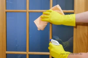 Close-up of hands wearing yellow gloves cleaning a wooden framed glass door using a spray bottle and cloth – representing detailed glass and surface cleaning.