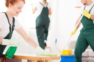 Smiling female cleaner wiping a table with spray bottle while team members clean windows and mop the floor in the background – representing professional room cleaning by Dirt2Tidy.