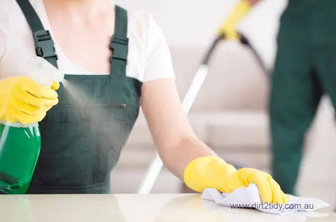 Close-up of a cleaner in uniform spraying green cleaning solution and wiping a white surface – with another cleaner vacuuming in the background, showcasing teamwork.