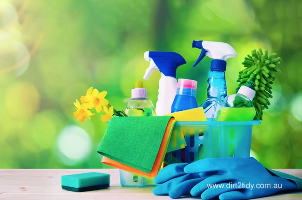 Colorful set of eco friendly cleaning products including spray bottles, microfiber cloths, sponges, and gloves arranged on a wooden table with a green outdoor background – promoting natural and safe cleaning solutions.
