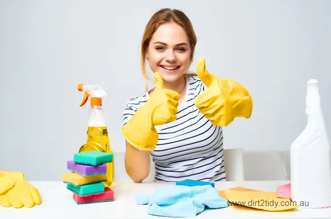 Smiling woman wearing yellow gloves giving thumbs up with colorful sponges, spray bottle, and cloths on the table – representing satisfaction with cleaning results.


