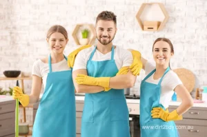 Smiling team of three professional cleaners wearing blue aprons and yellow gloves standing confidently in a modern kitchen – showcasing Dirt2Tidy’s expert staff.