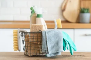 Basket filled with cleaning brushes, gloves, cloth, and sprays on a kitchen bench.