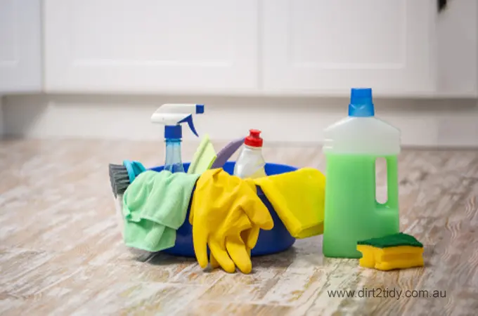 Assorted cleaning products and gloves placed on a wooden floor near white cabinets
