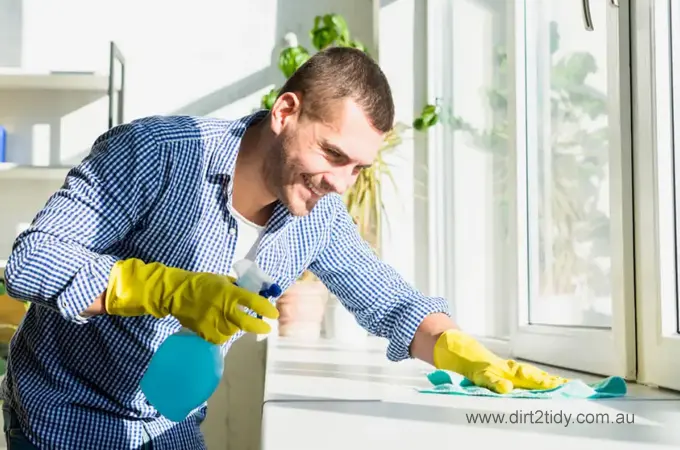 Smiling man in gloves using spray bottle and cloth to clean a sunny window sill.