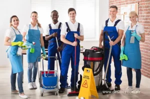 Group of professional cleaners in blue uniforms smiling and standing with cleaning tools, mop bucket, vacuum, and a caution wet floor sign – representing a reliable and prepared cleaning crew.