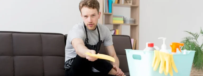 A man sitting on a sofa holding a paper while cleaning supplies are placed nearby, symbolising end-of-lease cleaning preparation.