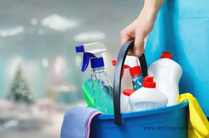 Hand holding a blue bucket filled with cleaning sprays and bottles.