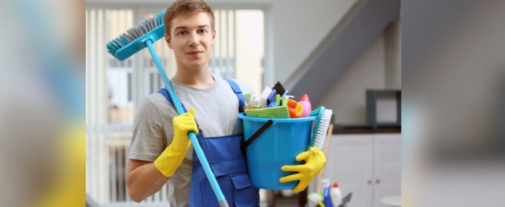 Young male cleaner wearing gloves and apron, holding a mop and a bucket filled with various cleaning supplies.