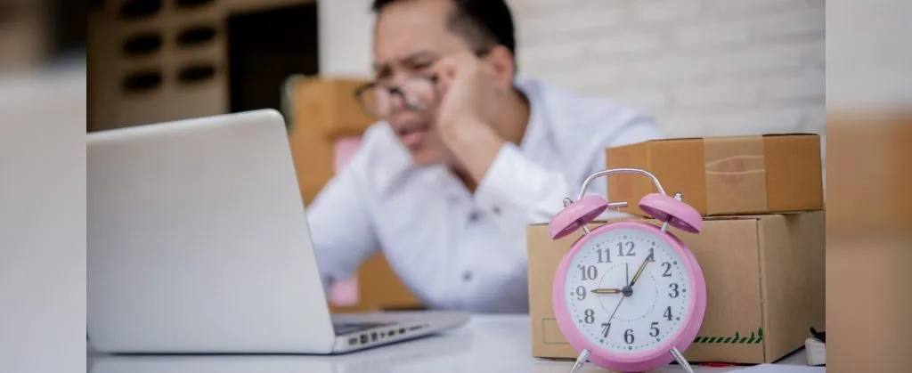 Frustrated man working on a laptop with moving boxes and a pink clock in the foreground, indicating time pressure.